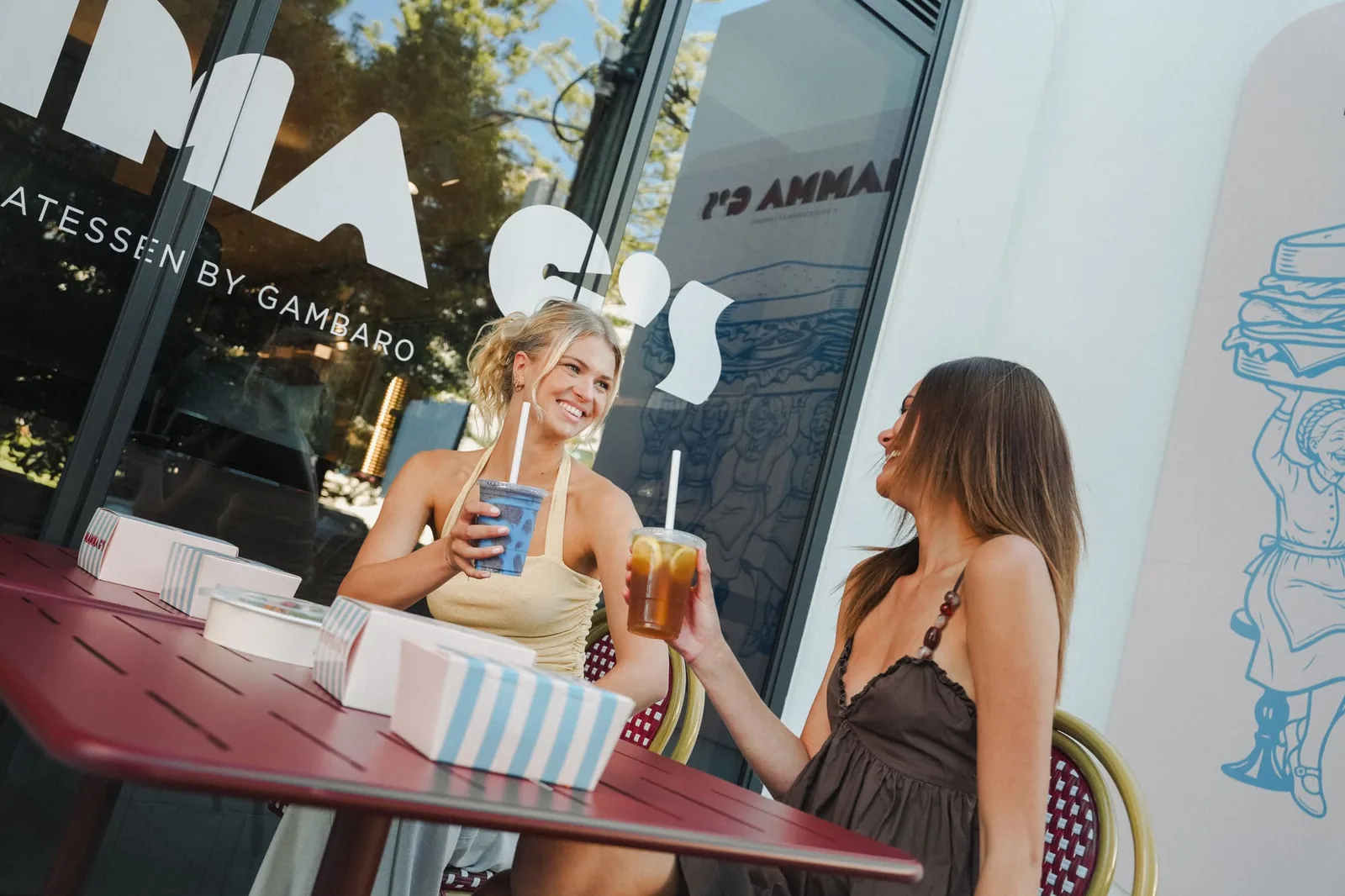 Two friends sitting at a red outdoor table outside Mamma G's, with the deli's signage and sandwich mural behind them